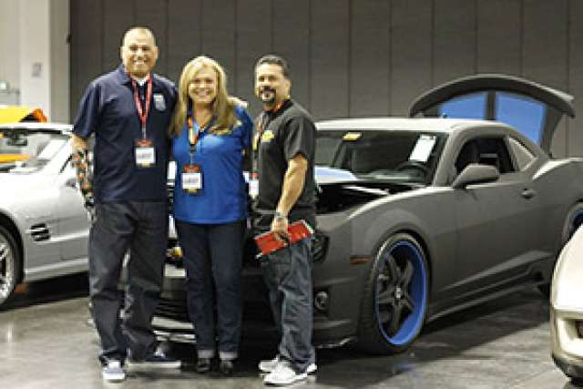Three people pose in front of a modified car at a car exhibition.