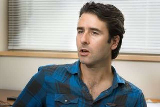 Man in a blue plaid shirt sitting in an office setting, facing slightly away.