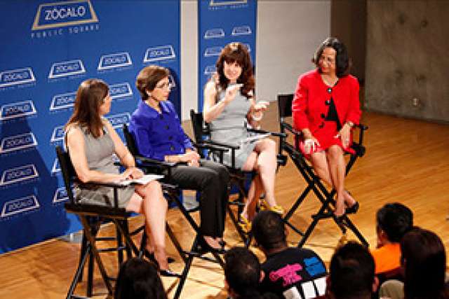 Four women discuss on stage at a public forum, with an audience in view.