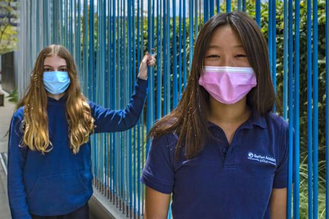 Two young girls outdoors, wearing masks, standing near a blue fence.