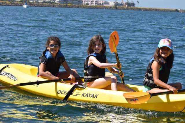 Three girls in life jackets paddle a yellow kayak together on calm water.