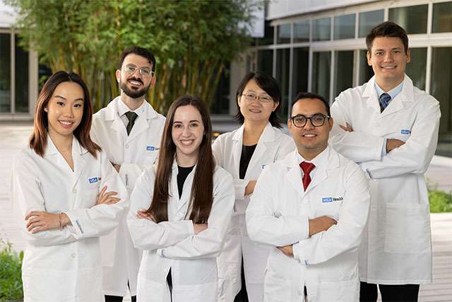 Six medical residents in white lab coats, standing outdoors with crossed arms.