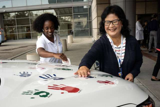 Two women smile while adding colorful handprints to the white hood of a car.