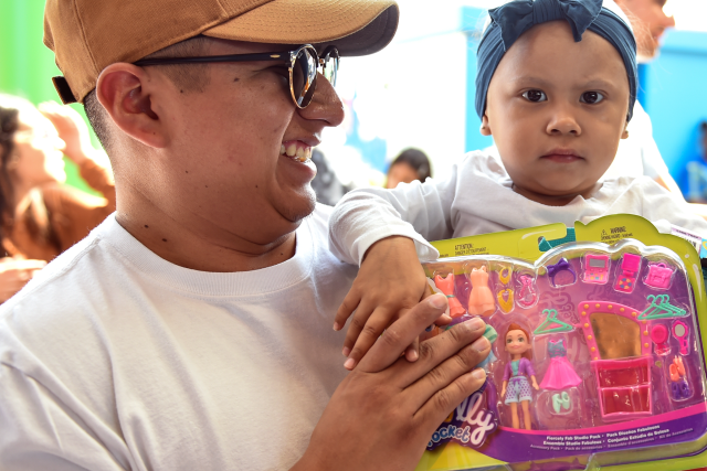 Smiling man holds a young girl with a blue headwrap and a new Polly Pocket toy set at a UCLA Health event.