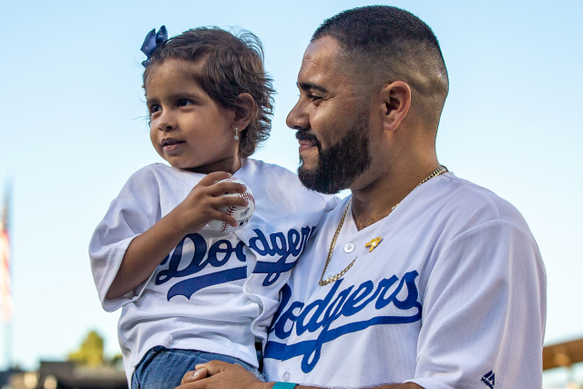 Father holds young daughter in Dodgers jersey on the field at Dodger Stadium, smiling during a special event.