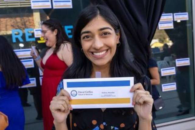  Nivedita Keshav smiles, holding up a white envelope with blue and yellow sections, standing in front of a window with similar envelopes.