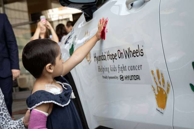 A young child with short dark hair, wearing a dark blue dress, places a red handprint on a white car door with "Hyundai Hope On Wheels" branding.