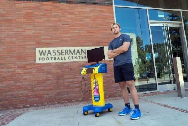Hayden standing beside a small yellow wheeled cart in front of Wasserman Football Centre sign