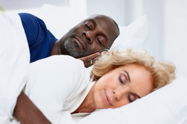 A couple, an African American man and a Caucasian woman, are sleeping peacefully in a white bed with their heads on a pillow.