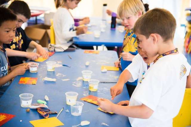 Children sit at a table doing crafts with cups, tools, and colorful paper during an indoor activity.