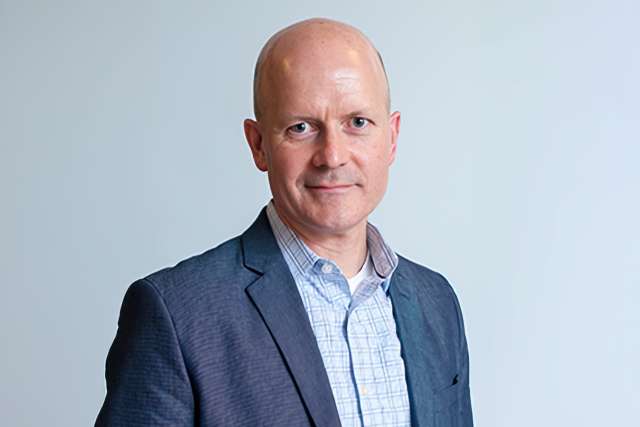 Dr. Keith Flahert in navy blazer and checked shirt against a light gray background.