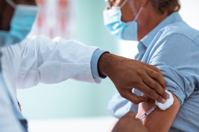 A medical professional, wearing a face mask and white coat, administers a vaccine to a patient's arm. The patient is also wearing a face mask.