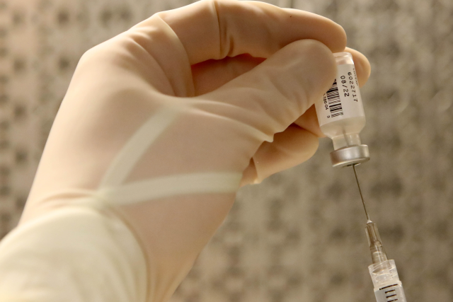 Someone wearing gloves and holding a vial of medicine, while puncturing it with a needle to withdraw medication.