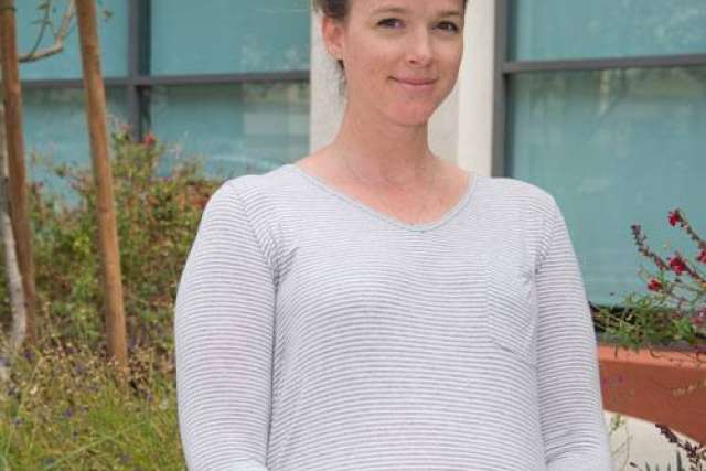Person wearing a light gray striped long-sleeve shirt standing outdoors near plants and a building with large windows.