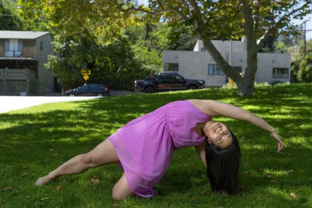 Janet Lo in a purple dress bends backward gracefully, one knee on the grass, with trees and houses in the background.
