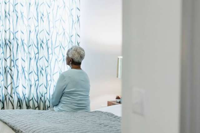 Older person seated on bed facing patterned curtains