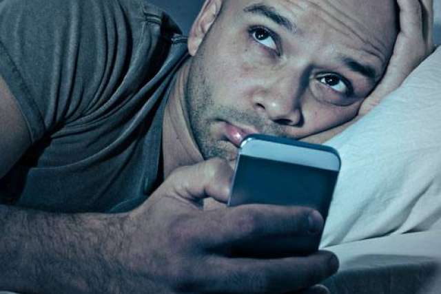 Person lying in bed holding a smartphone close, wearing a short-sleeve shirt with white bedding visible.