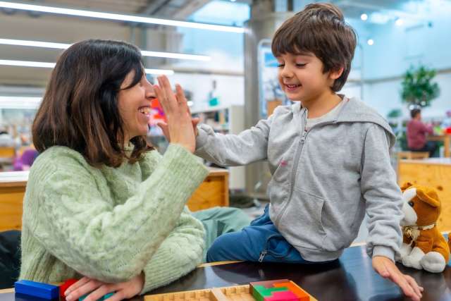 Girl holds wooden block to her eye like a toy camera; boy smiles and touches it