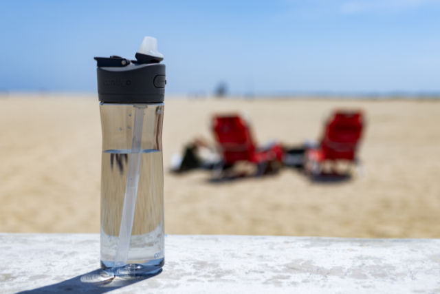 Plastic water bottle with cap and straw on a beach, with sand and some beach chairs in the background.