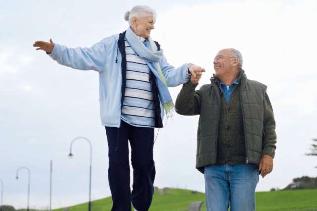 Two people outdoors, one balancing on a low ledge while holding hands with the other standing nearby on a grassy path.