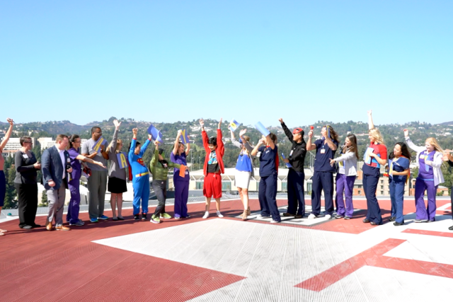 UCLA Health staff cheer on a rooftop helipad wearing capes and holding flags, celebrating as healthcare superheroes.