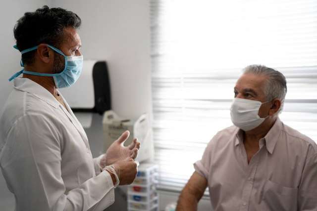 A healthcare professional wearing gloves and a mask speaks with a seated patient in a clinical setting near a window.