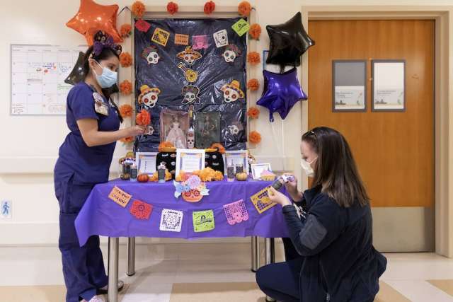Decorated table with colorful papel picado, flowers, balloons, and photos for a Day of the Dead display in a hallway.