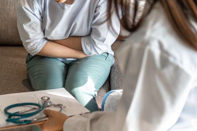 Person clutching lower abdomen during clinic visit; clinician with stethoscope visible.