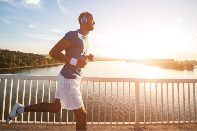 a man wearing white shorts, blue shirt, and headphones, running across the bridge as the sun sets.
