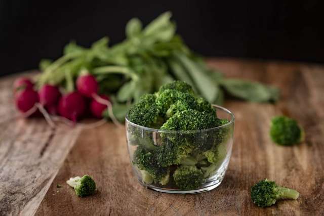 A bowl of fresh broccoli florets sits on a wooden surface, with red radishes and green leafy vegetables blurred in the background.