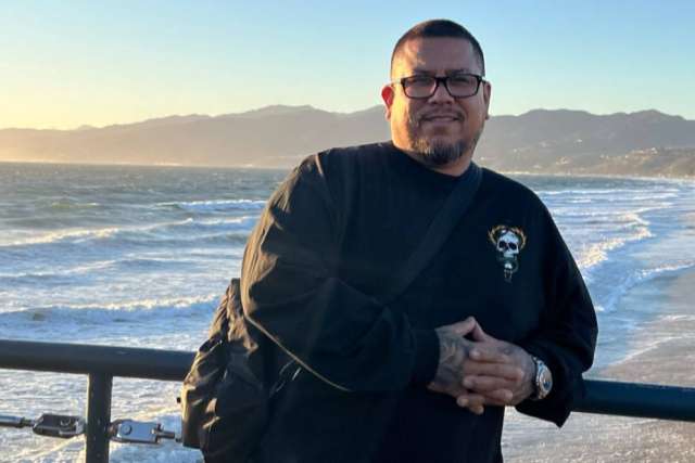 Person standing on a beachside railing with ocean and mountains in the background.