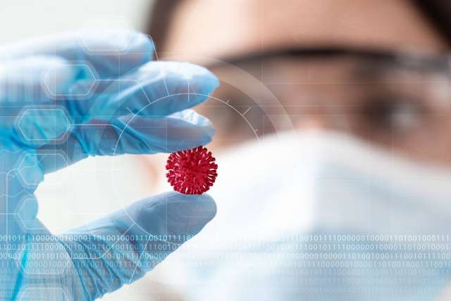 Scientist in mask and gloves holds a small red virus model, with faint digital graphics overlaying the image.