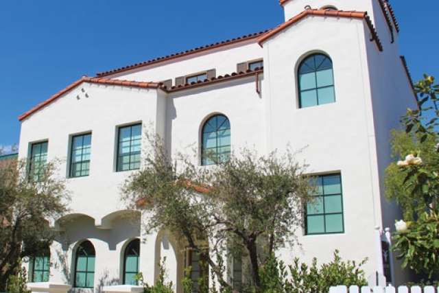White stucco building with multiple arched windows and terracotta roof tiles, surrounded by trees and greenery under a clear blue sky.