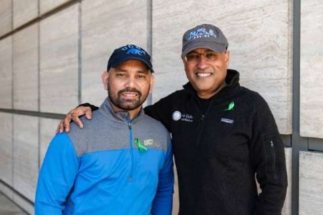 Two people wearing UCLA-branded caps and jackets with green awareness ribbons, standing side by side against a light stone wall