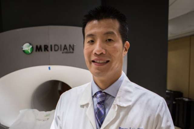 Person wearing a white lab coat and patterned tie standing in front of an MRIdian medical imaging machine in a clinical setting.