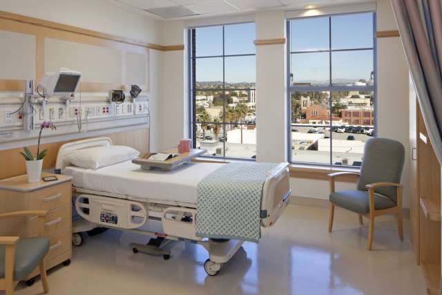 A bright, modern hospital room with a patient bed, medical equipment, a side chair, and two large windows offering a sunny view of a city landscape.