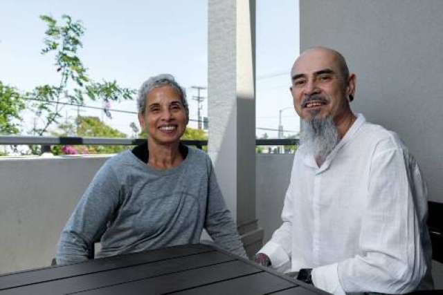 A man with a goatee, Felix Baltazar, and his wife, Peggy, a woman with gray hair, smile while sitting together at an outdoor table.