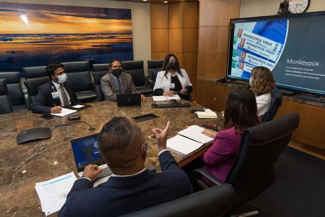People in business attire sit around a conference table while a presentation slide is displayed on a large screen.