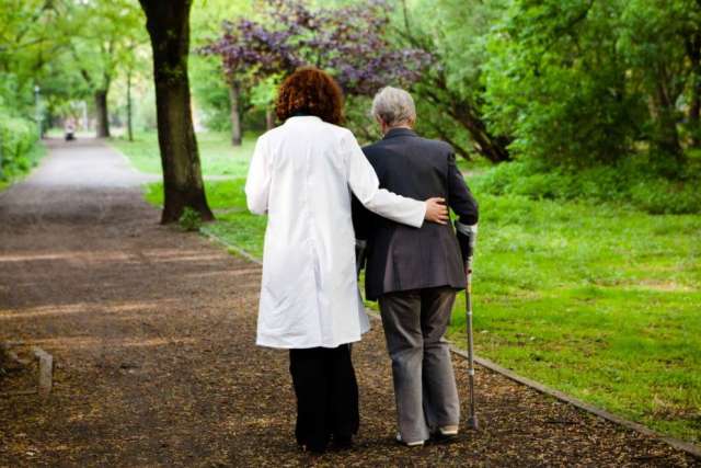 Caregiver in white coat supporting elderly person on park path
