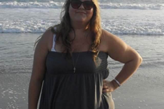 A smiling woman wearing sunglasses and a dark strapless sundress stands on a sandy beach with the ocean waves breaking behind her.