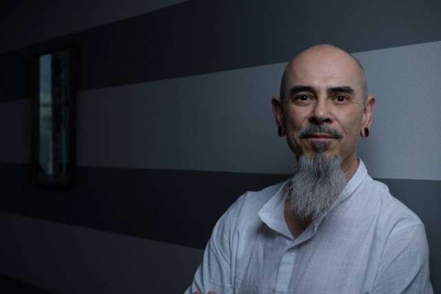 A professional headshot of Felix Baltazar, a bald man with a long gray goatee, looking confidently at the camera against a striped wall.