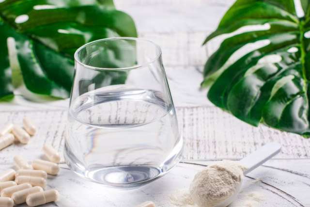 Glass of water with white capsules and powdered supplement on a white surface, green leaves in background.