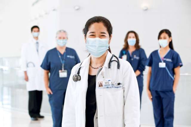 Group of masked healthcare workers, with a smiling female doctor in a white coat and stethoscope standing in front.