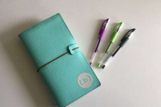 A closed teal journal with a white logo and elastic band lies next to three colorful pens (purple, green, and black) on a white surface.