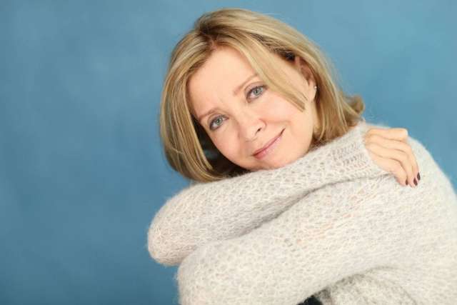 Rondi Charleston in a light sweater, hugging her knees against a blue background.