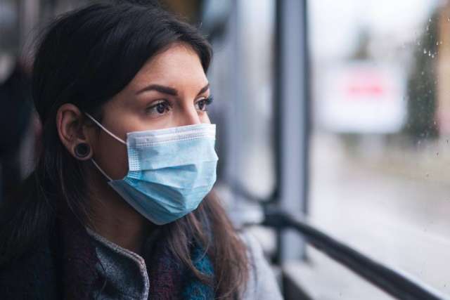 A young woman wearing a disposable light blue surgical face mask looks thoughtfully out a window while riding public transportation.
