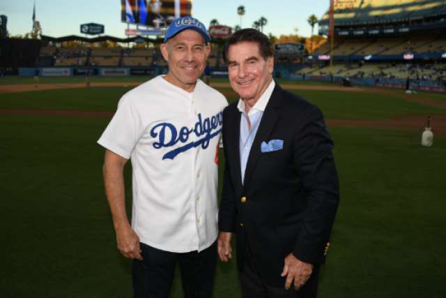 Dr. Mark S. Litwin in a Dodgers jersey and UCLA cap stands on a baseball field next to a man in a navy suit and light blue pocket square.