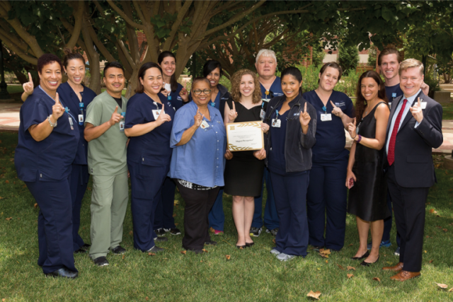 A diverse group of medical staff in scrubs stands outdoors, pointing upwards and celebrating, with a woman holding a recognition plaque.