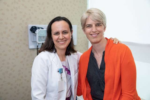  Lori Jordan, in an orange cardigan, and her doctor, in a white coat, stand close together, smiling at the camera in an exam room.