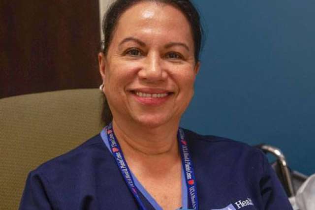 A person wearing a navy blue scrub top and a lanyard with text, seated indoors near a chair and a blue wall.